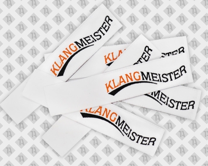 Gewebte Labels mit Schriftzug „KLANGMEISTER“ in Schwarz und Orange auf Weiß
