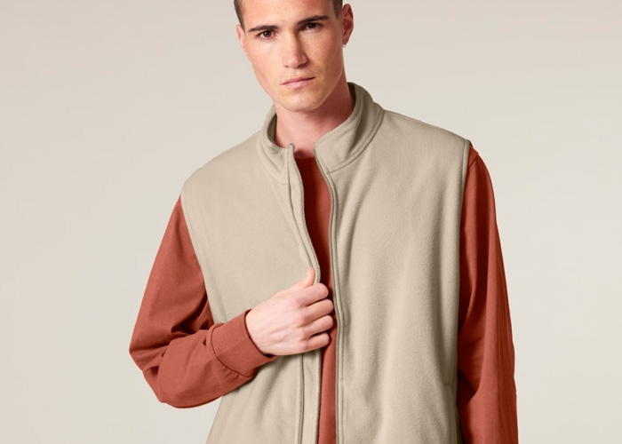 Ein junger Mann trägt eine beige Fleece-Weste über einem rostfarbenen Sweatshirt und hält sie vorne leicht fest.