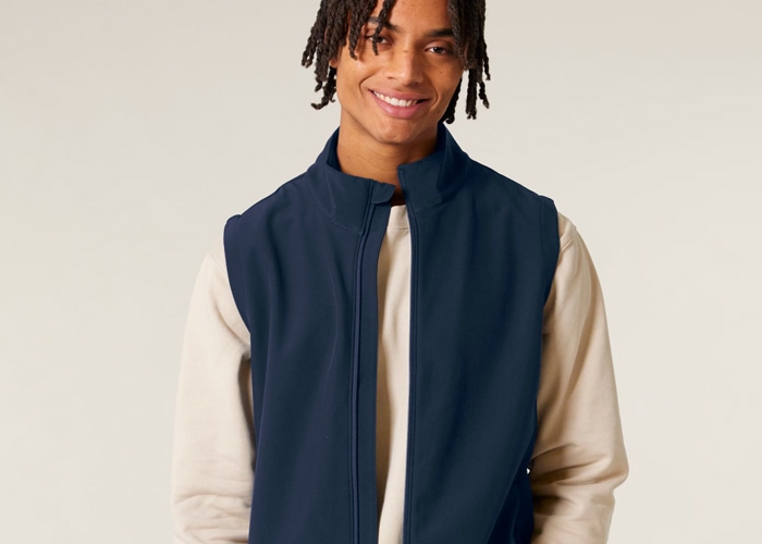 Ein lächelnder Mann trägt eine dunkelblaue Softshell-Weste über einem hellen Sweatshirt vor neutralem Hintergrund.