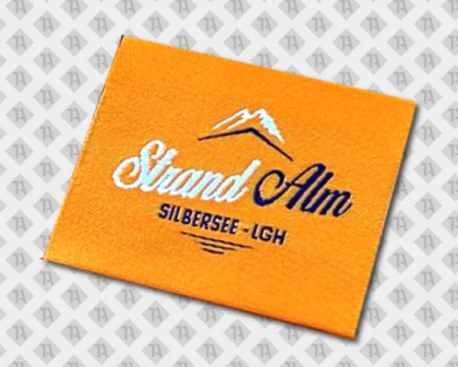 Gewebtes Label „Strand Alm Silbersee-LGH“ auf orangefarbenem Grund
