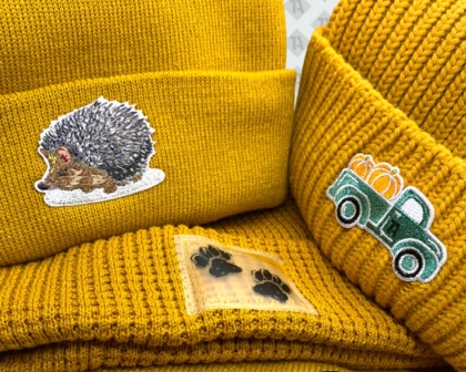 Drei senfgelbe Beanies mit gestickten Patches: Igel, Truck mit Kürbissen und Pfotenabdruck.